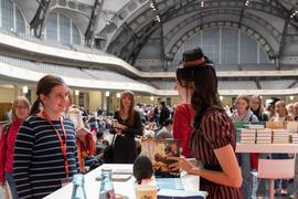 Autorinnengespräch am Büchertisch: Eine Autorin spricht mit einer Besucherin; im Hintergrund eine große Messehalle mit Menschen.