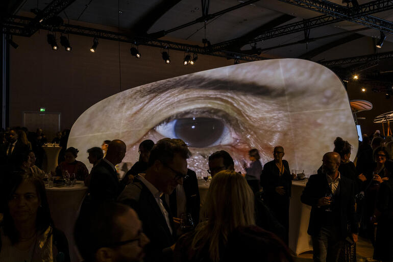 Menschen bei einem Empfang an Stehtischen in einem großen Saaal. Im Hintergrund eine Videoprojektion eines Auges.