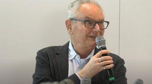 Ein Mann sitzt an einem Tisch und hält ein Mikrofon in der rechten Hand. Er spricht zum Publikum und trägt eine Brille.