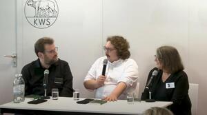 Zwei Männer und eine Frau sitzen nebeneinander an einem Tisch. Der Mann in der Mitte spricht in ein Mikrofon, dass er in der rechten Hand hält. Er schaut zu dem Mann auf der linken Seite. Dieser schaut zu dem Mann, der gerade spricht. Die Frau auf der rechten Seite schaut ebenfalls zu dem Mann in der Mitte. Alle tragen eine Brille.