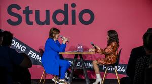 Zwei Frauen sitzen auf einer Bühne an einem Tisch und unterhalten sich. 