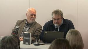 Zwei Männer sitzen nebeneinander an einem Tisch und schauen auf einen Laptop, der auf dem Tisch steht. Der Mann auf der rechten Seite spricht in ein Mikrofon. Auf dem Tisch steht ein Buch mit dem Titel "Vom Auswildern der Gespenster".