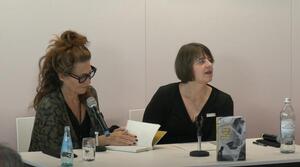 Zwei Frauen sitzen nebeneinander an einem Tisch. Die Frau auf der linken Seite trägt eine Brille und liest aus einem Buch vor, das aufgeschlagen vor ihr liegt. Die Frau auf der rechten Seite schaut zum Publikum. Auf dem Tisch steht ein Buch mit dem Titel "beat vor der eins". 