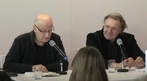 Zwei Männer sitzen nebeneinander an einem Tisch. Der Mann auf der linken Seite trägt eine Brille und liest aus einem Buch vor, das vor ihm auf dem Tisch liegt. Der Mann auf der rechten Seite schaut zum Buch.