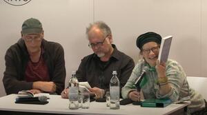 Zwei Männer und eine Frau sitzen nebeneinander an einem Tisch. Die Frau sitzt auf der rechten Seite, lächelt und hält ein Buch hoch. Der Mann in der Mitte schaut auf ein Buch, welches er in der Hand hält. Der Mann auf der linken Seite schaut auf den Tisch. Alle Personen tragen eine Brille.