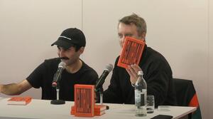 Zwei Männer sitzen nebeneinander an einem Tisch. Der Mann auf der rechten Seite schaut ins Publikum und hält mit der linken Hand ein Buch mit dem Titel "Gloria!" hoch. Der Mann auf der linken Seite lächelt und schaut ebenfalls ins Publikum.