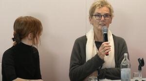 Zwei Frauen sitzen an einem Tisch. Die linke Frau schaut zur rechten Frau. Die Frau auf der rechten Seite trägt eine Brille und spricht in ein Mikrofon. Dabei schaut sie ins Publikum.