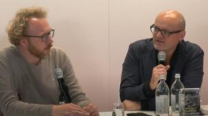 Zwei Männer sitzen nebeneinander an einem Tisch, beide tragen eine Brille. Der Mann auf der rechten Seit hält ein Mikrofon in der Hand und spricht, dabei schaut er leicht zum linken Bildrand. Der Mann auf der linken Seite schaut zum rechten Bildrand. Auf dem Tisch steht ein Buch mit dem Titel "Aus unseren Feuern".