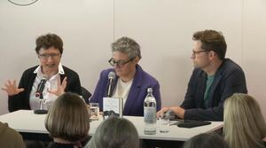 Zwei Frauen und ein Mann sitzen nebeneinander an einem Tisch, alle tragen eine Brille. Die Frau auf der linken Seite spricht zum Publikum und gestikuliert mit ihren Händen. Die Frau in der Mitte schreibt etwas auf ein Papier. Der Mann auf der rechten Seite schaut die Frau auf der linken Seite an.