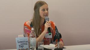 Eine Frau sitzt an einem Tisch und liest aus einem Buch vor, das aufgeschlagen vor ihr liegt. In ihrer rechten Hand hält sie ein Mikrofon. Auf dem Tisch steht ein Buch mit dem Titel "Lorettas letzter Trip". 