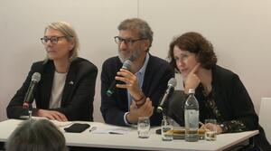 Zwei Frauen und ein Mann sitzen nebeneinander an einem Tisch. Der Mann in der Mitte hält ein Mikrofon in der Hand. Alle Personen schauen ins Publikum.