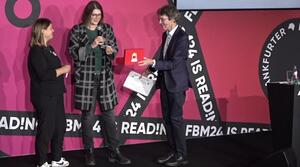 Zwei Frauen und ein Mann stehen nebeneinander auf einer Bühne. Der Mann steht rechts und hält eine rote Box fest. Die Frau in der Mitte zieht einen Zettel aus der Box und spricht in ein Mikrofon. 