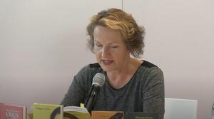 Eine Frau sitzt an einem Tisch und liest aus einem Buch vor, das sie aufgeschlagen vor sich hält. Vor ihr stehen vier Bücher auf dem Tisch.