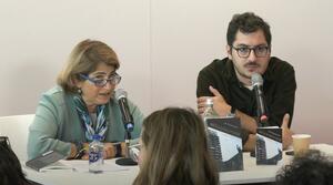 Eine Frau und ein Mann sitzen nebeneinander an einem Tisch. Beide Personen tragen eine Brille und schauen zum Publikum. Die Frau sitzt auf der linken Seite, der Mann sitzt auf der rechten Seite.