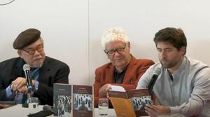 Drei Männer sitzen nebeneinander an einem Tisch. Der Mann auf der rechten Seite liest aus einem Buch vor, das er aufgeschlagen vor sich hält. Die beiden anderen Männer tragen eine Brille und schauen ihn an. Auf dem Tisch stehen zwei Bücher mit den Titel "Last Call Manila" und "Killing Time in a Warm Place".