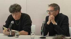 Zwei Männer sitzen nebeneinander an einem Tisch. Der Mann auf der linken Seite liest aus einem Buch vor. Der Mann auf der rechten Seite trägt eine Brille, hat seine Hände ineinander verschränkt und schaut ins Publikum.