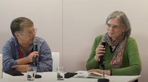 Zwei Frauen sitzen nebeneinander an einem Tisch. Beide Frauen tragen eine Brille und halten in ihrer rechten Hand ein Mikrofon. Die Frau auf der rechten Seite spricht zum Publikum, die Frau auf der linken Seite schaut zu ihr. Vor der Frau der rechten Seite liegt ein aufgeschlagenes Buch auf dem Tisch.