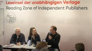 Zwei Männer und eine Frau sitzen nebeneinander an einem Tisch, die Frau sitzt in der Mitte. Alle tragen eine Brille. Der Mann auf der rechten Seite spricht in ein Mikrofon und schaut zu den anderen Personen. Die beiden anderen Personen schauen zu ihm, die Frau lächelt.