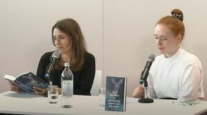 Zwei Frauen sitzen nebeneinander an einem Tisch. Die Frau auf der linken Seite liest aus einem Buch vor, das sie aufgeschlagen vor sich hält. Die Frau auf der rechten Seite schaut auf den Tisch. Auf dem Tisch steht ein Buch mit dem Titel "Gratulieren müsst ihr mir nicht" von Lily Polansky.