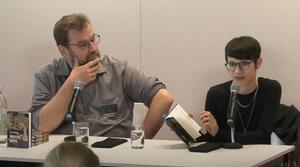 Ein Mann und eine Frau sitzen nebeneinander an einem Tisch. Der Mann auf der linken Seite schaut zur Frau auf der rechten Seite und hat seine rechte Hand an das Kinn gelegt. Die Frau der linken Seite spricht in ein Mikrofon und blättert in ihrem Buch. Beide Personen tragen eine Brille. Auf dem Tisch liegen drei Bücher, davor steht ein Buch mit dem Titel "Mariens Käfer" von Lisa Kränzler.