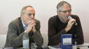 Zwei Männer sitzen nebeneinander an einem Tisch. Der Mann auf der rechten Seite trägt eine Brille und spricht in ein Mikrofon, das er in seiner rechten Hand hält. Der Mann auf der linken Seite hat seine Hände verschränkt und an das Kinn gelegt und schaut zum Publikum. Auf dem Tisch steht ein Buch mit dem Titel "Am Tag zuvor, am Tag danach".