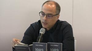 Ein Mann sitzt an einem Tisch und liest aus einem Buch vor, das er  aufgeschlagen vor sich hält. Auf dem Tisch stehen zwei Bücher mit dem Titel "1983".