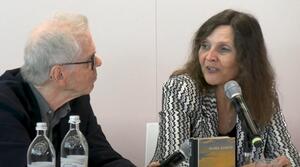 Ein Mann und eine Frau sitzen nebeneinander an einem Tisch. Die Frau auf der rechten Seite spricht in ein Mikrofon und schaut zu dem Mann auf der linken Seite. Dieser trägt eine Brille und schaut die Frau an. Auf dem Tisch steht ein Buch von Maike Albath.