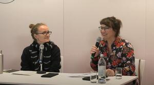 Zwei Frauen sitzen nebeneinander an einem Tisch. Beide tragen eine Brille. Die Frau auf der rechten Seite hält ein Mikrofon in der rechten Hand und spricht zum Publikum. Die Frau auf der linken Seite schaut die andere Frau an und lächelt.