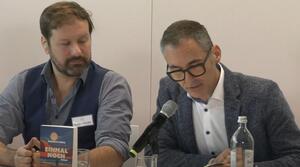 Zwei Männer sitzen nebeneinander an einem Tisch. Der Mann auf der rechten Seite trägt eine Brille und liest aus einem Buch vor, das aufgeschlagen vor ihm liegt. Der Mann auf der linken Seite schaut zum aufgeschlagenen Buch. Auf dem Tisch steht ein Buch mit dem Titel "Einmal noch".