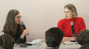 Zwei Frauen sitzen nebeneinander an einem Tisch. Die Frau auf der linken Seite spricht in ein Mikrofon und schaut die Frau auf der rechten Seite an. Die Frau auf der rechten Seite blickt zur Frau auf der linken Seite.