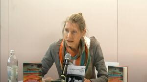 Eine Frau sitzt an einem Tisch und liest aus einem Buch vor, das aufgeschlagen vor ihr liegt. Sie schaut zum Publikum. Auf dem Tisch stehen zwei Bücher mit dem Titel "Fremder Champagner" und ein Flyer mit der Aufschrift "Der Ilse Schwepcke Preis".