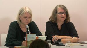 Zwei Frauen sitzen nebeneinander an einem Tisch. Die linke Frau liest aus dem aufgeschlagenen Buch vor ihr vor. Die Frau auf der rechten Seite trägt eine Brille und schaut zum Publikum.