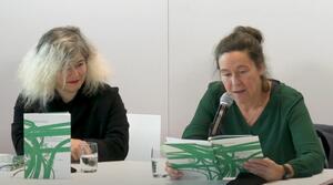 Zwei Frauen sitzen nebeneinander an einem Tisch. Die Frau auf der rechten Seite spricht in ein Mikrofon und liest aus einem Buch vor, das sie aufgeschlagen vor sich hält. Die Frau auf der linken Seite schaut zu dem aufgeschlagenen Buch. Auf dem Tisch steht eine weitere Ausgabe des Buches mit dem Titel "Höllenfahrt & Entenstaat".