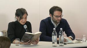 Ein Mann und eine Frau sitzen nebeneinander an einem Tisch. Die Frau auf der linken Seite liest aus einem Buch vor, das sie aufgeschlagen vor sich hält. Der Mann auf der rechten Seite trägt eine Brille und blättert in einem Buch, das vor ihm liegt.