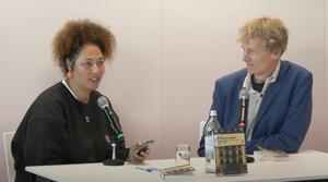 Zwei Personen sitzen nebeneinander an einem Tisch. Die Frau auf der linken Seite spricht in ein Mikrofon und schaut zum Publikum. Der Mann auf der rechten Seite schaut zur Frau.