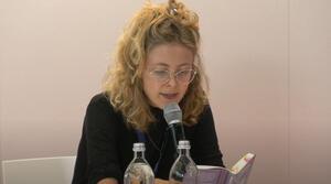 Eine Frau sitzt an einem Tisch und liest aus einem Buch vor, das sie aufgeschlagen vor sich hält. Sie trägt eine Brille.