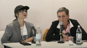 Zwei Frauen sitzen nebeneinander an einem Tisch. Die Frau auf der rechten Seite spricht in ein Mikrofon. Die Frau auf der linken Seite schaut zum Publikum.