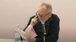 Ein Mann sitzt an einem Tisch und liest aus dem aufgeschlagenen Buch vor sich. Er trägt eine Brille fasst sich mit dem rechten Zeigefinger an den Kopf.