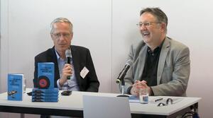 Zwei Männer sitzen an einem Tisch, beide tragen eine Brille. Der Mann auf der linken Seit hält in seiner rechten Hand ein Mikrofon und spricht, dabei schaut er zum Publikum. Der Mann auf der rechten Seite schaut zum Publikum und lacht.