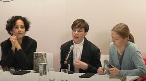 Drei Frauen sitzen an einem Tisch. Die Frau in der Mitte spricht in ein Mikrofon und schaut zum Publikum. Die Frau auf der rechten Seite schreibt etwas auf ein Blatt Papier.