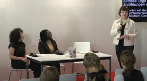 Zwei Frauen sitzen nebeneinander an einem Tisch. Eine weitere Frau steht rechts neben dem Tisch, spricht in ein Mikrofon und schaut zum Publikum. Die beiden Frauen am Tisch lächeln und schauen zur Frau neben dem Tisch.