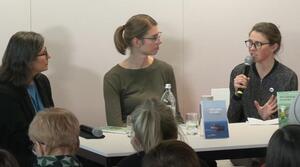 Drei Frauen sitzen nebeneinander an einem Tisch. Die Frau auf der rechten Seite hält ein Mikrofon in ihrer rechten Hand und spricht zu den anderen beiden, dabei gestikuliert sie mit ihrer linken Hand. Die beiden anderen Frauen schauen zu ihr. Alle tragen eine Brille. Auf dem Tisch stehen zwei Bücher mit den Titeln "Une loge en mer" und "Die Congierce ist auf See".