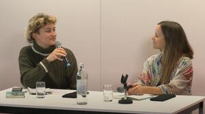 Zwei Frauen sitzen nebeneinander an einem Tisch und schauen sich lächelnd an. Die Frau auf der linken Seite hält ein Mikrofon in ihrer rechten Hand und spricht. Vor ihr lieg ein Notizblock und ein Buch auf dem Tisch.