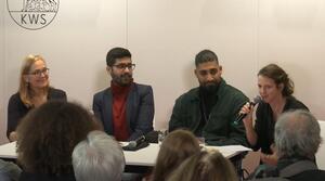 Zwei Frauen und zwei Männer sitzen nebeneinander an einem Tisch. Die Frauen sitzen links und rechts außen. Die Frau auf der rechten Seite spricht in ein Mikrofon und schaut in die Menge. Die anderen drei Personen lächeln und schauen zu der Frau mit dem Mikrofon.