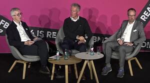Drei Männer sitzen nebeneinander auf einer Bühne. Der linke Mann redet zu den anderen beiden Männern.