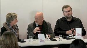 Zwei Männer und eine Frau sitzen nebeneinander an einem Tisch. Der Mann in der Mitte hält ein Mikrofon in der rechten Hand und spricht zur Frau auf der linken Seite. Der Mann auf der rechten Seite trägt eine Brille und schaut zum Publikum. Auf dem Tisch liegen und stehen mehrere Bücher mit dem Titel "Die Möglichkeit einer Micky Maus".