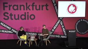 Zwei Frauen sitzen nebeneinander auf der Bühne. Die linke Frau schaut runter und die rechte Frau schaut ins Publikum. Rechts von den Frauen steht ein Rednerpult und im Hintergrund zeigt ein Bildschirm Logo und Veranstaltungstitel.