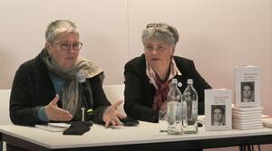 Zwei Frauen sitzen nebeneinander an einem Tisch. Die linke Frau spricht zum Publikum und gestikuliert mit ihren Händen. Die Frau auf der rechten Seite schaut leicht zum linken Bildrand. Auf dem Tisch liegen und stehen mehrere Exemplare eines Buches mit dem Titel "Ein Mann ohne Titel".