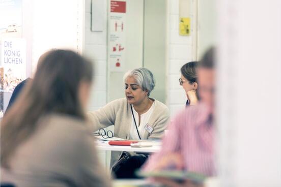 Im Vordergrund zwei Menschen am Tisch sitzen im Gespräch, verschwommen sichtbar, im Hintergrund zwei Frauen im Gespräch 