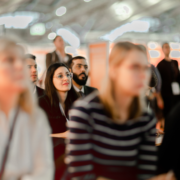 Foto von Menschen, die gemeinsam in einer Messehalle sitzend einem Vortrag lauschen. Die Blicke alle in dieselbe Richtung gerichtet. 